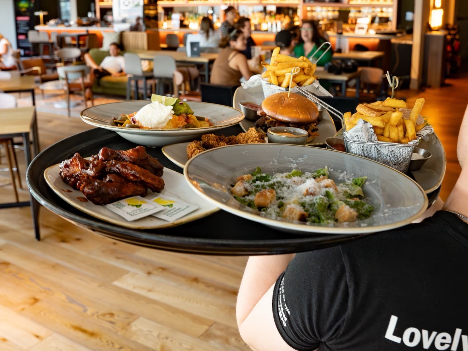 A waiter carrying a tray of barbecue ribs, nachos, a burger with fries and a Caesar salad at Caddi Club Wimbledon