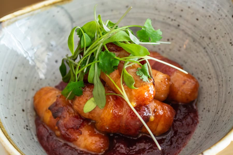Pigs in blankets, sausages wrapped in bacon with a berry glaze and fresh herbs, in a bowl